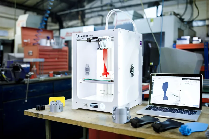 Drukarka 3D FDM drukująca czerwoną część, obok laptop z podglądem modelu w slicerze i gotowe wydruki na stole warsztatowym