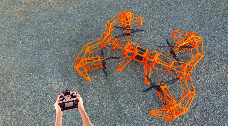 Duży pomarańczowy dron z ażurową ramą wydrukowaną w 3D i czterema śmigłami w kratowych osłonach, stojący na asfalcie; obok dłonie trzymające aparaturę RC
