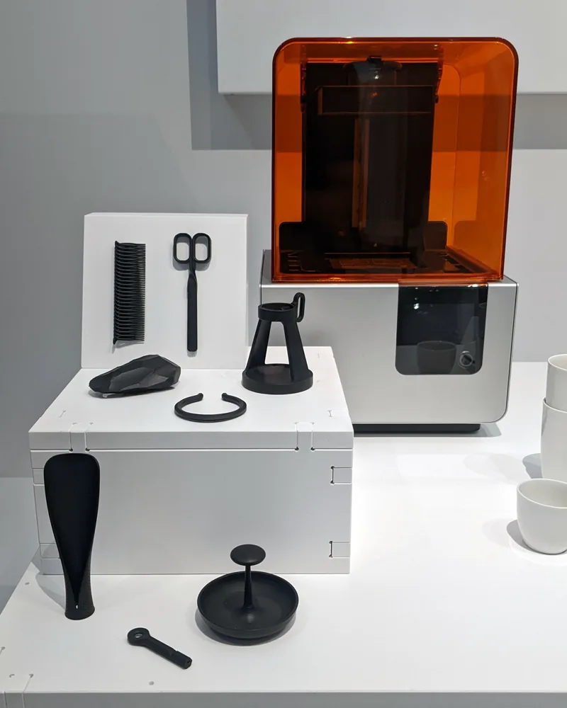 Drukarka żywiczna SLA z pomarańczową pokrywą, a obok niej czarne przedmioty wydrukowane w technologii 3D, na wystawie muzealnej