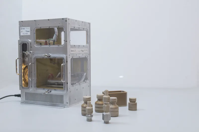 Zamknięta drukarka 3D z metalową obudową i oknami, obok wydrukowane części z beżowego tworzywa